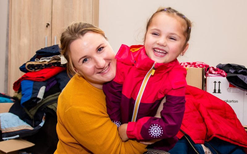 A young woman and a girl crouch amid boxes of coats and clothes, smiling at the camera
