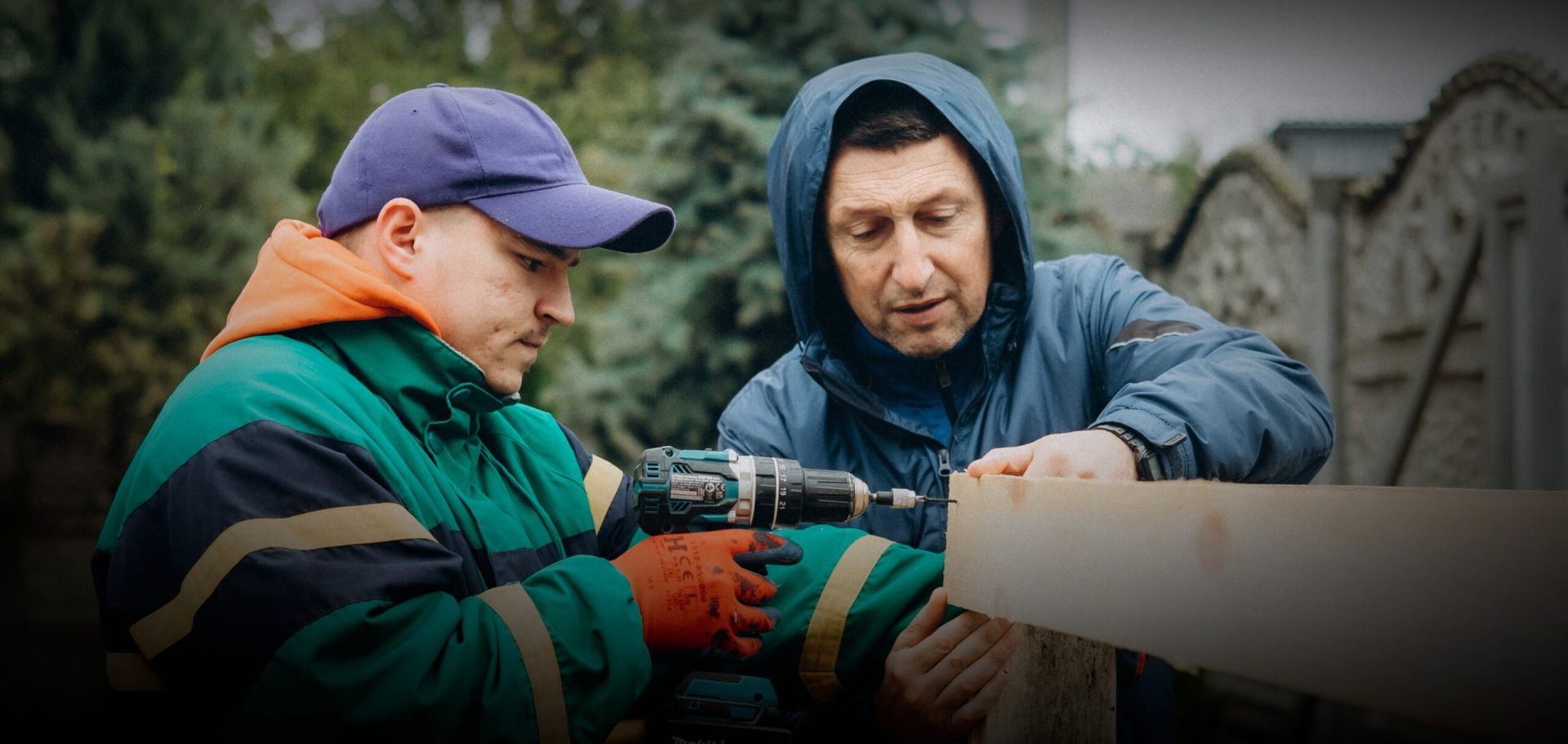 A young man and a middle-aged man use power tools outside on a wooden framework